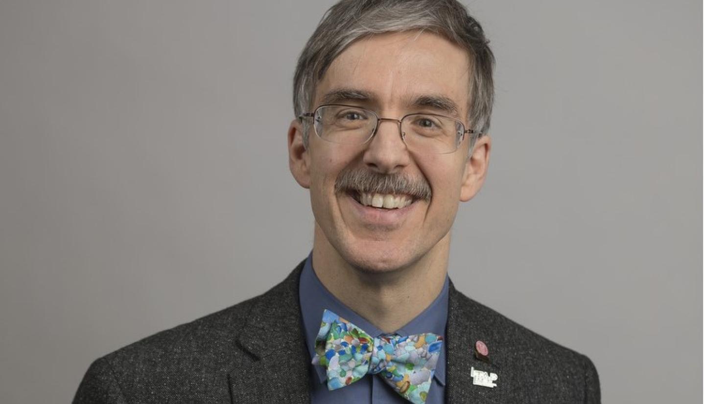 A man wearing a blue shirt, gray suit jacket with a bowtie and glasses smiling for a photo