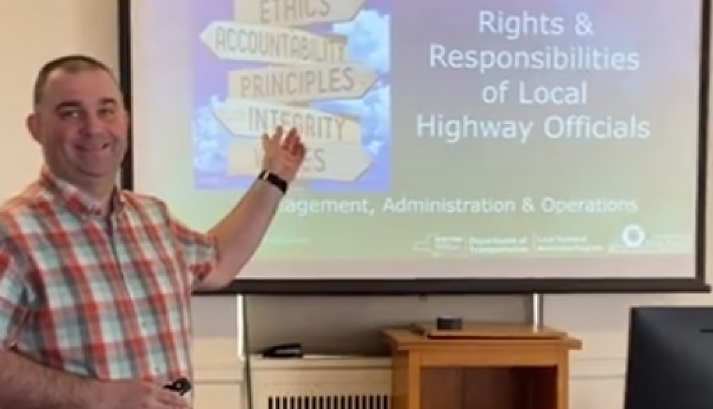 A man wearing a red, blue and white plaid shirt. standing in front of a screen giving a presentation. 