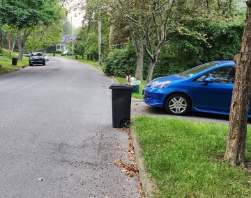 A residential street with a black trash bin in the road and a blue mini van trying to pull out of a driveway