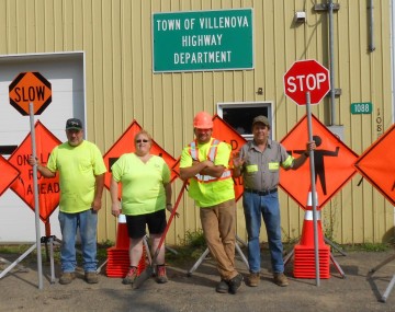 Road workers stand among several road work signs.